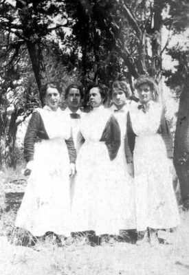 A photograph of five of the Harvey Girls in full uniform. No location is specified, but they are standing in front a grove of trees.