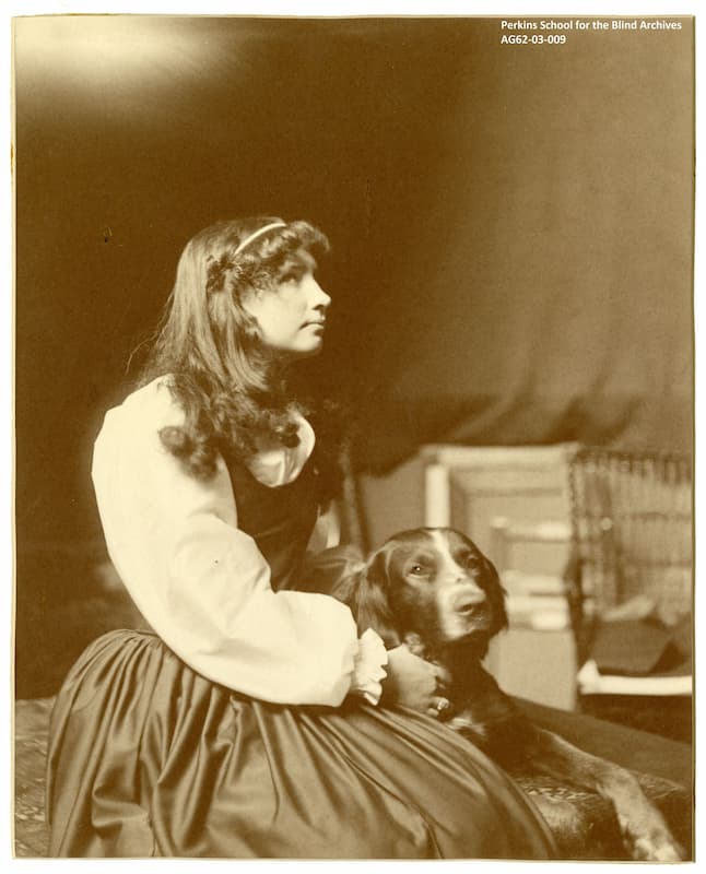Helen Keller as an adolescent, sitting next to another of the family dogs. She is dressed in a white blouse and dark full skirt. Her hair is long.