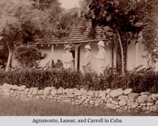 This is an of-the-area photograph of Drs. Agramonte, Lazear, and Carroll. They are standing and talking under a shelter. Each is wearing a light-colored uniform and pith hat-style helmet.