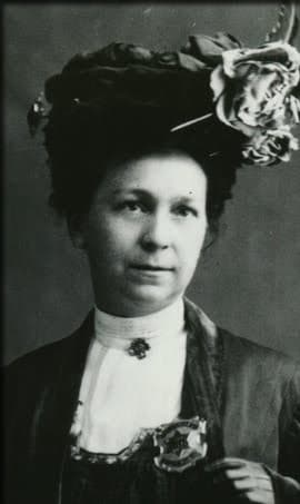 Alice S. Wells in a professional photograph. She is dressed up and wears a hat with a flower atop it. She also wears her police badge.