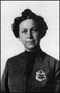 Alice Stebbins Wells in a dark jacket and wearing her badge. Her hair is in a top knot. She wears no hat.