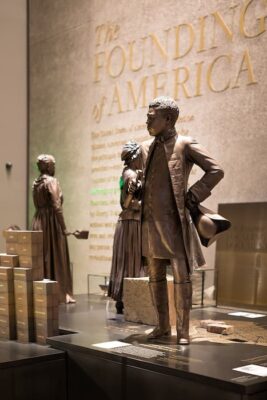 Three bronze statues of historical figures, including Banneker, stand in front of a wall engraved with The Founding of America. Stacks of gold bars and plaques are displayed nearby in the museum exhibit.