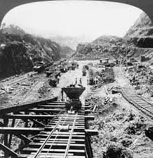 This is a contemporary black-and-white photo of the canal being built. A rail line is visible on the right side and some type of construction equipment (maybe a small dumpster?) is in the center of the photo.
