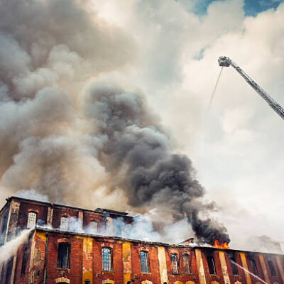 A burning brick building with great grey plumes of fire rising from the rooftop.