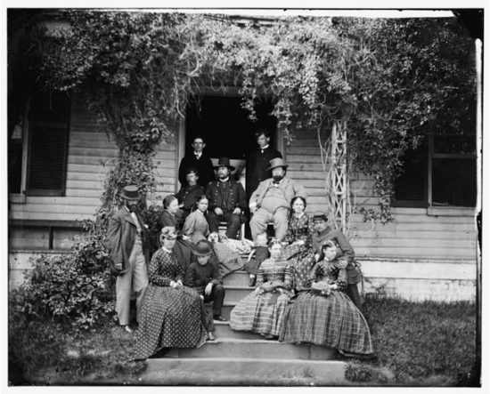 A photograph of the Ingalls family. The Rufus Ingalls sits in a chair on the porch; his dog is at this feet. The rest of family surrounds them.