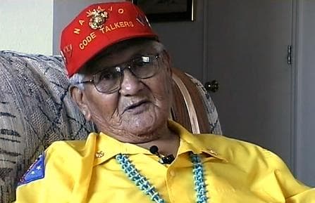 Photo of Chester Nez telling his story. He wears a red billed cap with "Navajo Code Talkers" on it.