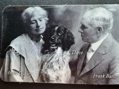 Black and white photo of Annie Oakley, a man labeled Frank Butler, and a dog labeled Dave. Annie and Frank face each other with the dog in between them, all posed against a plain backdrop.