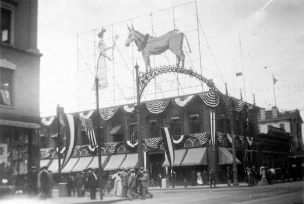 Black-and-white photo of a corner building decorated with patriotic bunting, flags, and a large sign featuring a donkey above the roof. People gather on the sidewalk below, suggesting a lively scene at the Security 1908 Convention.