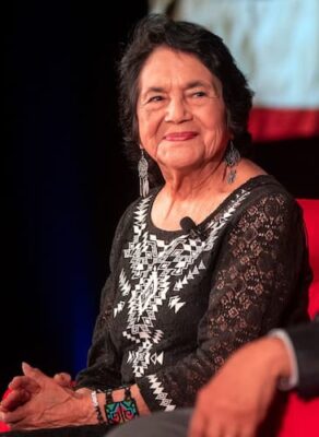 A recent color photo of Dolores Huerta. She wears a decorative black top and silver dangling earrings.