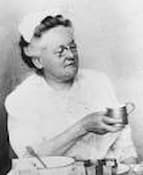 black and white photo of Fannie Farmer demonstrating in a cooking class. She is holding a measuring cup.