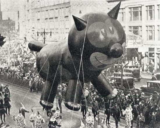 This a black-and-white photo of the Felix the Cat balloon making its way down Broadway. There are clowns serving as balloon handlers, and the years is probably late 1920s.