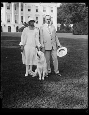 Grace and Calvin Coolidge outside the White House. They are dressed for summer weather and one of the white collies is with them.