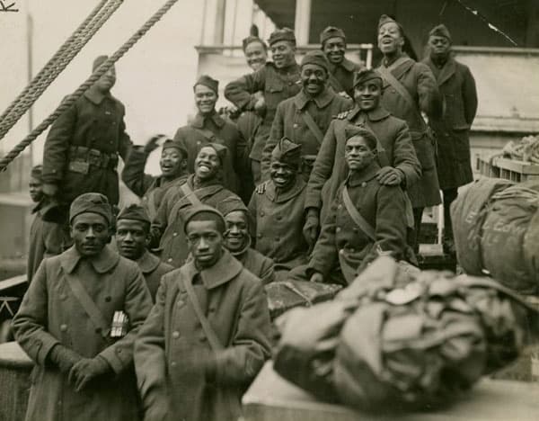 A black-and-white photograph of about a dozen Harlem Hellfighters mugging for the camera on shipboard.