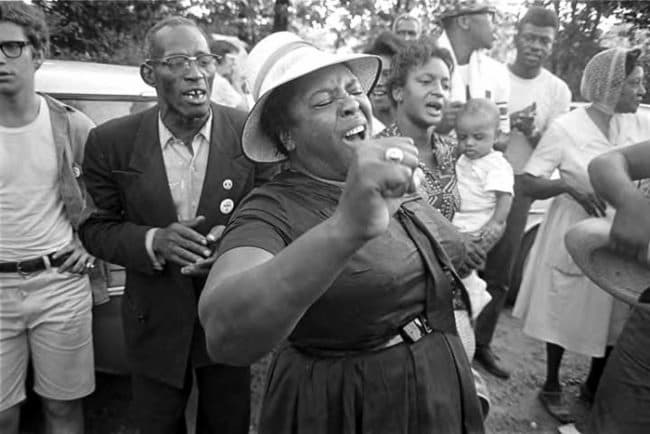 Hamer stands in the midst of a group of people. She has on a dress and hat, and the holds a microphone in her right hand. She is clearly belting out a song.