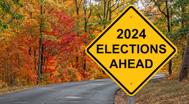 Photographer Jim Vallee  places a yellow "warning sign" of "Elections Ahead" on what appears to be a beauiful country road in the autumn. The leaves are green, red, and gold.