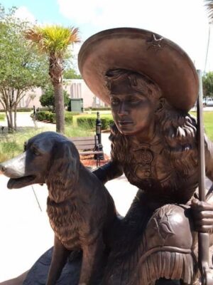 A bronze statue of a woman, Annie Oakley, wearing a cowboy hat kneeling beside Dave, with trees and a building in the background on a sunny day.