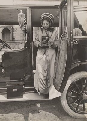This is a promotional photo of an elegantly dressed young woman in hat and coat, stepping out of a turn-of-the-century automobile. She aims a Kodak camera.