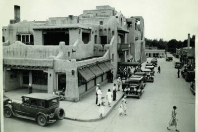 This is a black-and-white photo of the exterior of the La Fonda Hotel in Santa Fe. People are milling around on the street, and there are cars from the late 1930s or early '40s.