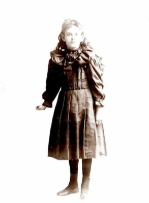 This is a photograph of Mary Jane Arnold as a child. Her hair is long and curly, and seh is dressed in a dark dress and tights.