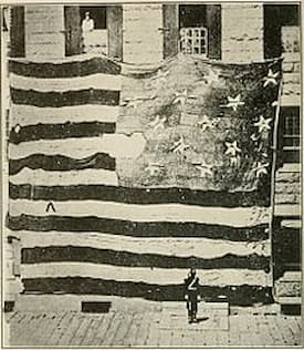 A large, old U.S. flag from Fort McHenry with 15 stars and 15 stripes is displayed on the side of a stone building. One person stands on a balcony above the flag and another below, emphasizing the impressive size of the historic banner.