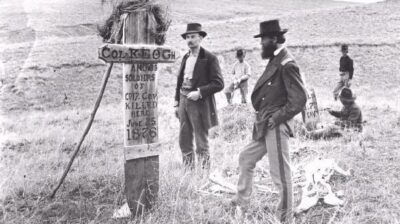 A black-and-white photo showing men who have arrived after the battle is over. They stand, trying to make sense of what has happened. One man in the background is painting names on markers to note who died and where they were.
