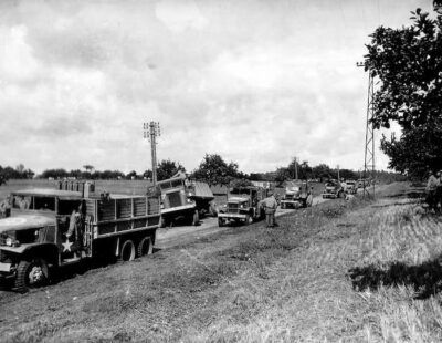 A black and white photo of a truck convoy. One truck is pulled over and stalled. The other 5 visible trucks are making their way past the stalled one.