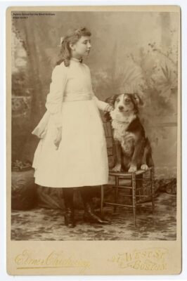 Helen Keller as a young girl standing by one of the dogs she had as a child. The dog sits on a stool. Helen, wearing a white dress, stands beside the dog with one hand on his bak.