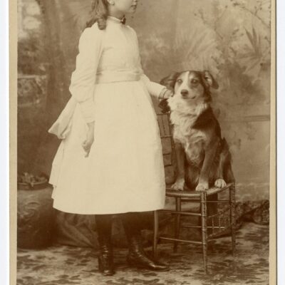 Helen Keller as a girl standing by one of her dogs who sits on a stool.