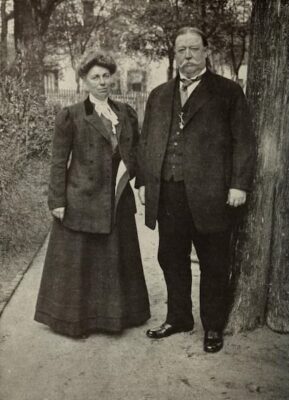 Helen and William Taft in formal early 1900s attire stand together on a path outdoors, next to a tree, with trees, a fence, and what appears to be Taft’s cow in the background.