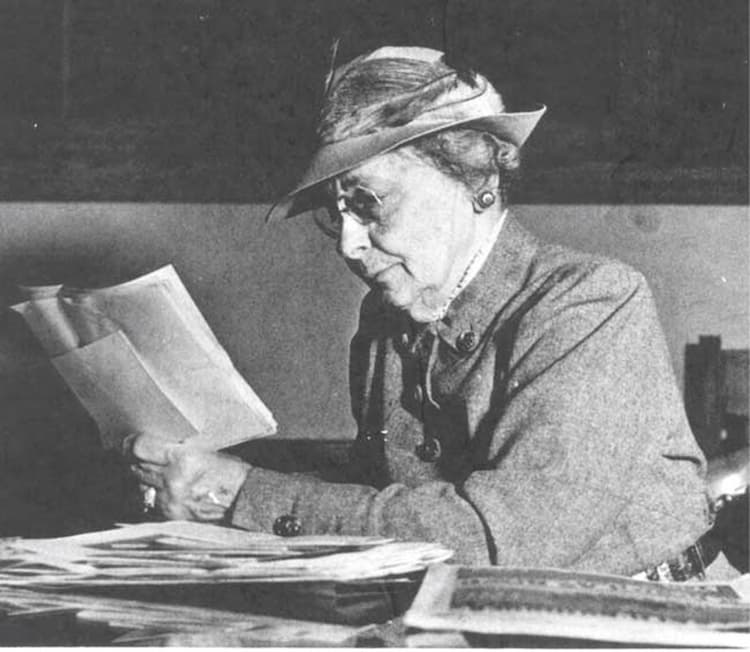 An elderly Rose Knox sits at a table with her work piled in front of her. She is reading what may be a map of sales territories. She is dressed neatly in a suit with a fashionable hat.