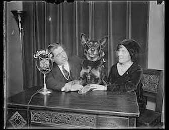 A delightful press photo ofSenator Schall and his wife, who are to be interviewed by Columbia Radio. Lux stands between them with both paws on the table, seemingly knowing he is the star of the photo. 