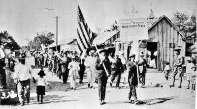 A post card of a parade in Webb County. An American flag leads ooff the procession.