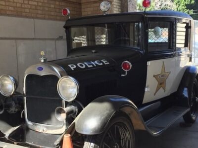 This is a photograph of a police car. It like dates to the early 1940s. It's box-like closed car with a star on the door an "POLICE" written across the car hood.