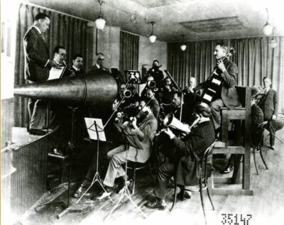 The photograph shows a small band. Everyone is crowded around the recording horn. The men are all dressed in suits, and one fellow stands on the stage and seems to be in charge
