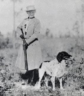 Black-and-white photo of Annie Oakley in a long coat and hat holding a rifle, standing outdoors beside a medium-sized dog with a white and dark coat.