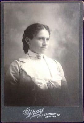 A black-and-white portrait of a woman in Victorian-era clothing, with braided hair, posed in front of a cloudy backdrop. The bottom of the photo says Gray 1090 Tremont St. Boston.