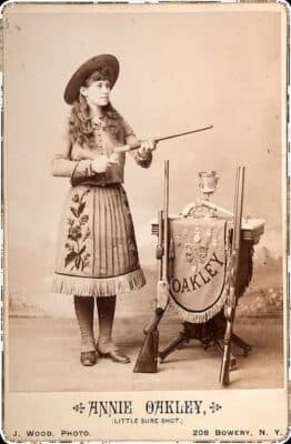 Sepia photo of Annie Oakley wearing a dress and hat, holding a rifle. Next to her is a table with trophies, rifles, and a fringed banner reading Oakley. As the legendary sharpshooter of Buffalo Bill's Wild West Show, she stands poised and confident. Text below reads Annie Oakley, Little Sure Shot.