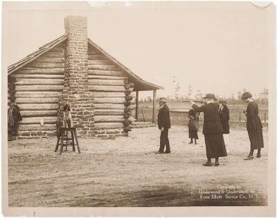 Five people, dressed in early 20th-century clothing, stand outside a log cabin with a stone chimney. One person appears to be aiming a camera at a child sitting on a stool in front of the cabin.