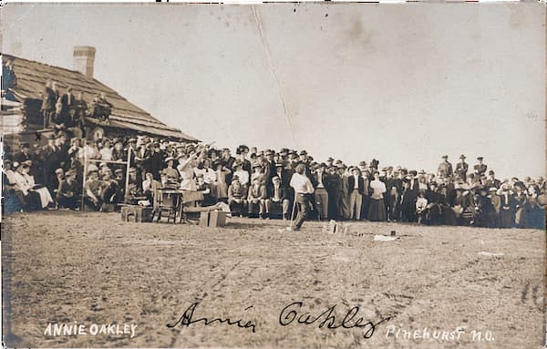 Black-and-white photo of a large crowd gathered outdoors, watching a woman perform. “Annie Oakley” and “Pinehurst, N.C.” are written on the image. A wooden building is visible in the background.