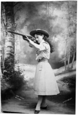 This is a posed shot of Annie Oakley shooting her rifle. She looks fully in command, standing with her left foot forward, wearing her hair long with a hat on top.