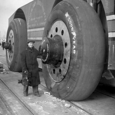 Man stands beside the Antarctic snow cruiser. Though the man is tall, the tire is huge--standing 10 feet high.