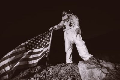 An astronaut in a spacesuit stands on a rocky surface, proudly holding a U.S. flag. The black and white scene captures the essence of a historic moon landing.
