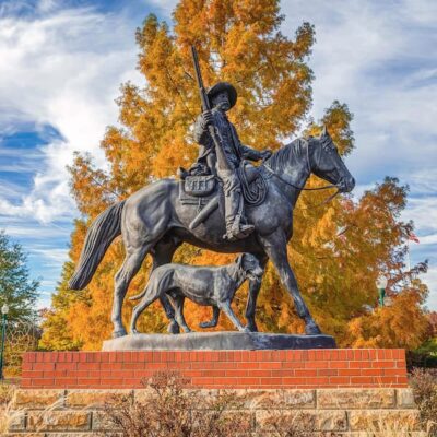 A stunning bronze sculpture of Bass Reeves on horseback. He holds his Winchester rifle in his right hand. A dog follows along beside him. The photograph was taken in the autumn, and the tree behind the statue is golden.