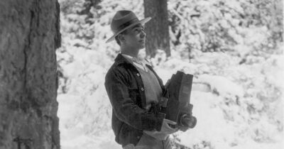 A black-and-white photo of George Wright in the field. He wears a park ranger hat and hold a notebook and camera.