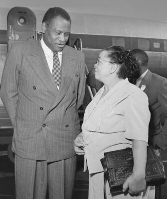 Paul Robeson wears a double-breasted pin-striped suit and stand on the airport tarmac where he talks to Charlotta Bass.  They are very focused on their conversation.