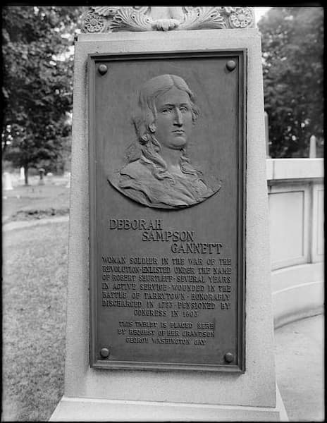 A bronze plaque on a stone monument honors Deborah Sampson, a courageous Female Soldier in the Revolutionary War, with her relief portrait and an inscription. Trees and part of a fence can be seen in the background.