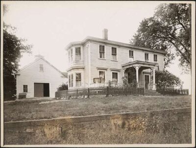 Historic black-and-white photo of a two-story wooden house with a porch, surrounded by a picket fence. Deborah Sampson home.