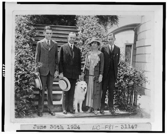 Calvin and Grace Coolidge are flanked by their two sons. All are in suits and ties. Grace wears a day dress. Prudence Prim stands with them.