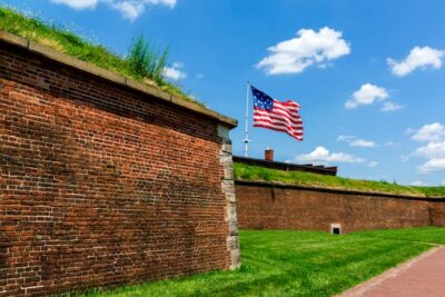 A large U.S. flag waves above the brick walls of historic Fort McHenry under a bright blue sky with scattered clouds and lush green grass in the foreground.