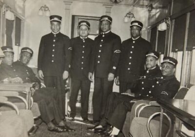 This is a black-and-white photograph of 8 Pullman porters in uniform. They are standing in a rail car; 4 are sitting, the otehr 4 are standing. The men seem cvurious about why the photo is being taken. Library of Congress